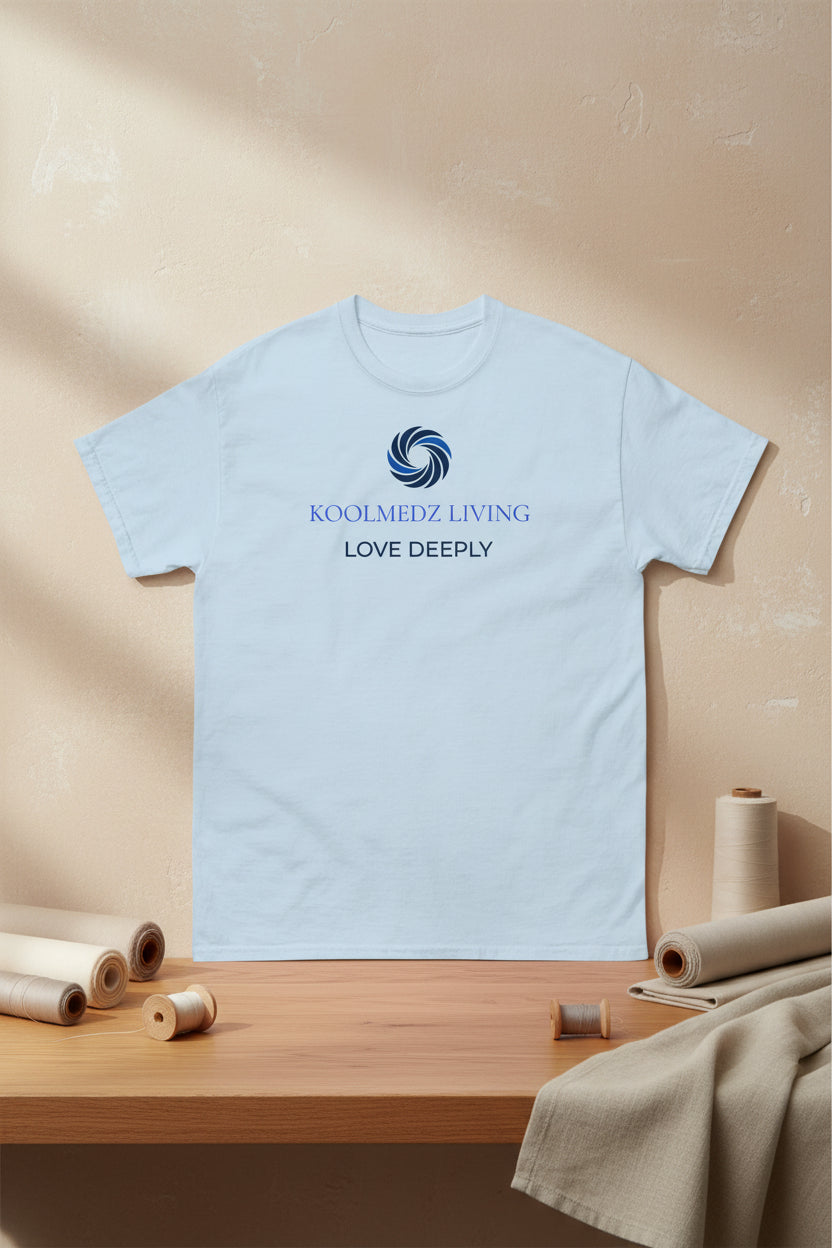 A light blue short-sleeve crewneck t-shirt with a blue and navy graphic on the chest featuring a swirl logo and the text 'KOOLMEDZ LIVING' and 'LOVE DEEPLY'.