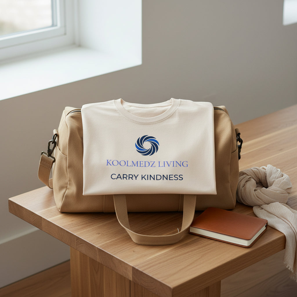 A light beige crewneck t-shirt with a blue and navy graphic on the chest featuring a swirl logo and the text 'KOOLMEDZ LIVING' and 'CARRY KINDNESS'.