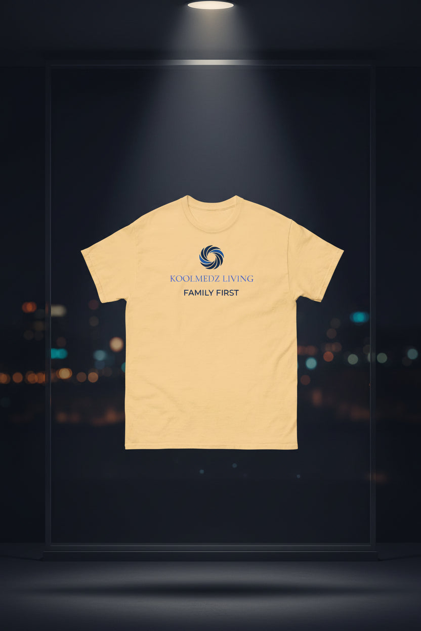 A light peach-beige crewneck t-shirt with a blue and navy graphic on the chest reading 'KOOLMEDZ LIVING' and 'FAMILY FIRST' with a swirl logo, displayed on a clean white background.